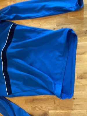 Blå Nike sporttröja - Blå långärmad Nike sporttröja med en mörkblå och vit rand över bröstet. Tillverkad i ett mjukt och stretchigt syntetmaterial som passar perfekt för träning och sport.