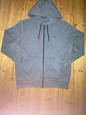 Ralph Lauren grå zip - Snygg grå hoodie med dragkedja och huva. Tröjan har svarta snören i huvan, två fickor framtill och en liten broderad logga på bröstet. Perfekt för en chill och avslappnad stil. Tillverkad i mjukt sweatshirtmaterial.