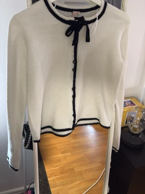 Vit kofta med svarta detaljer och rosett - Söt vit kofta med svarta kanter och dekorativ rosett vid halsen. Koftan har knappar framtill och långa ärmar. Passar perfekt till både jeans och kjol för en chic look.