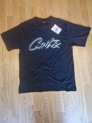 Svart cortiez tröja - Snygg svart t-shirt med stor vit grafisk logga på bröstet. Klassisk passform och rund halsringning. Perfekt till jeans eller shorts för en avslappnad streetwear-look.