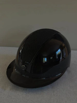Glossy glitter ridhjälm - Snygg svart ridhjälm med glansig finish och glitterdetaljer på ovansidan. Hjälmen har en klassisk form med solskärm och justerbart hakband i brunt läder. Perfekt för dig som vill ha både stil och säkerhet i stallet. Går att anpassa storleken 