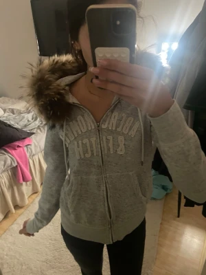 Grå hoodie med fuskpäls från Abercrombie - Mysig grå zip-hoodie från Abercrombie & Fitch med stor luva kantad av fuskpäls. Tröjan har dragkedja framtill, två fickor och tryckt logga över bröstet. Perfekt för kalla dagar och chill outfits.