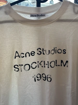 Vit Acne Studios - Vit t-shirt från Acne Studios med svart tryck framtill där det står 'Acne Studios STOCKHOLM 1996'. Klassisk rund halsringning och avslappnad passform. Perfekt till jeans eller shorts för en clean och stilren look.
