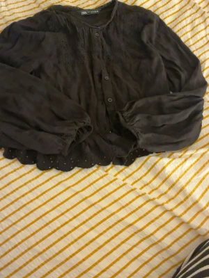 Svart blus med spetsdetaljer från Zara - Svart blus från Zara med långa ärmar, knappar framtill och fina spetsdetaljer vid halsringning och nederkant. Blusen har en lös passform och vågig kant längst ner. Perfekt för en trendig och avslappnad look.