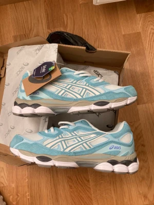  Nya Asics blåvita sneakers -  Nya Snygga sneakers från Asics i ljusblått och vitt med mesh och mockadetaljer. Skorna har en chunky sula i vitt och grått samt klassiska Asics-loggor på sidan. Perfekta för dig som gillar sportig och trendig stil.
