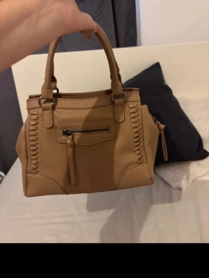 Beige handväska med detaljer - Snygg beige handväska i mjukt konstläder med två handtag och dekorativa sömmar på sidorna. Väskan har dragkedjefack både fram och bak samt en tofsdetalj på dragkedjan. Perfekt storlek för att få plats med det viktigaste.
