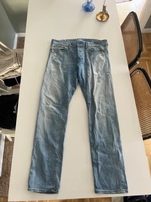 Ljusblå raka jeans med slitningar - Säljer ett par ljusblå jeans med raka ben och diskreta slitningar framtill. Jeansen har klassisk femficksdesign och är tillverkade i denim. Perfekta för en avslappnad och trendig look.