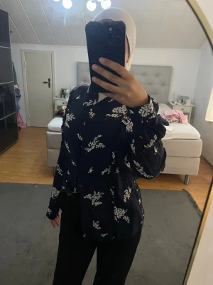 Mörkblå blommig blus med knappar - Snygg mörkblå blus med vita blommönster och knäppning framtill. Blusen har långa ärmar med breda manschetter och en avslappnad passform. Perfekt att styla med både jeans och kostymbyxor för en trendig look.