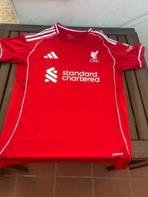 Liverpool röd matchtröja Adidas - Säljer en röd Liverpool matchtröja från Adidas med vita detaljer och klubbmärke på bröstet. Tröjan har rund hals, vita ränder på axlarna och tryckt sponsorlogga framtill. Tillverkad i lätt och ventilerande material, perfekt för fotboll eller träning.