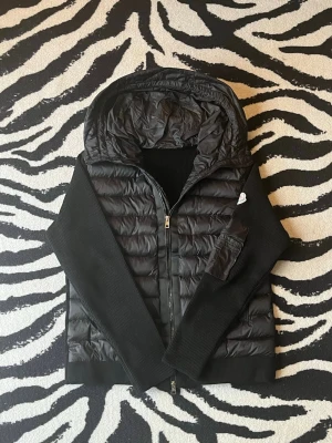 Moncler Cardigan - Snygg svart cardigan från Moncler med stickade ärmar och huva. Jackan har dragkedja framtill, ribbade muddar och en ficka med dragkedja på ärmen. Moncler-logga på vänster ärm. Perfekt för kalla vinterdagar eller till hösten. Pris kan diskuteras.