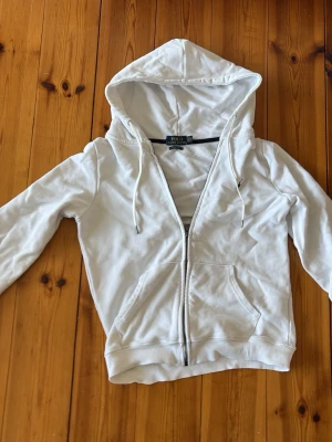 Vit hoodie från Polo Ralph Lauren - Snygg vit hoodie från Polo Ralph Lauren med dragkedja framtill, två fickor och justerbar huva med snören. Klassisk design med diskret logga på bröstet. Tillverkad i mjukt bomullsmaterial som är skönt att ha på sig.