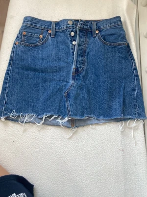 Blå jeanskjol från Levis - Säljer en klassisk blå jeanskjol i denim, 5 knappar framtill men bara en syns .Den har en fransig kant nedtill. Kjolen har fem fickor och bälteshällor. 
