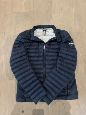 Mörkblå pufferjacka från Colmar - Snygg mörkblå pufferjacka från Colmar med quiltad design och snygg krage. Jackan har dragkedja framtill och en diskret logga på ärmen. Perfekt för kalla dagar och ger en stilren look. Tillverkad i lätt och värmande material. Passar perfekt till kalla sommardagar och till våren/hösten.