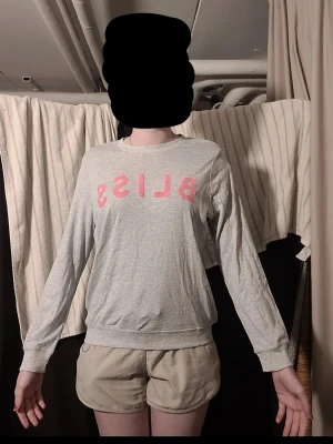 Grå sweatshirt med rosa text - Ljusgrå sweatshirt med rund halsringning och långa ärmar. På bröstet står det 'BLISS' i stora rosa bokstäver. Tröjan har ribbade muddar vid ärmslut och nederkant. Perfekt för en avslappnad och trendig look.