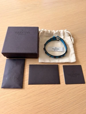 Valentino armband  - Valentino armband i en väldigt snyggt ljusblå färg med silvriga nitar, passar perfekt nu till sommaren! Självklart är det äkta! Köpt på NK för 3200:- (tyvärr inget kvitto) alla nitar finns kvar, otroligt bra skick. Har tecken på användning men fortfarande mycket kvar att ge! Alla tillbehör som följde med vid köp och som syns på bild medföljer!