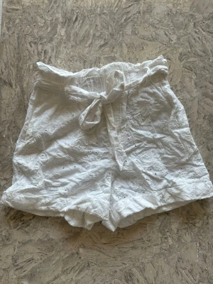 Vita broderade shorts med knyt från Zara  - Säljer ett par vita shorts med broderade blommor och snygg knyt i midjan. De är högmidjade och har en luftig passform, perfekta för varma dagar. Materialet känns som bomull och shortsen har fina detaljer med volangkant upptill.