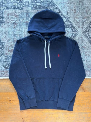 Mörkblå hoodie från Polo Ralph Lauren - Snygg mörkblå hoodie från Polo Ralph Lauren med vit dragsko och klassisk röd logga broderad på bröstet. Hoodien har känguruficka framtill och ribbade muddar vid ärmar och nederkant. Perfekt för en avslappnad och stilren look.