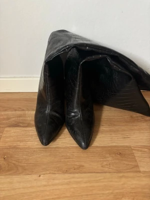 Höga stövlar - Säljer ett par svarta stövletter med hög klack och spetsig tå. Skorna har ett snyggt krokopräglat mönster. 