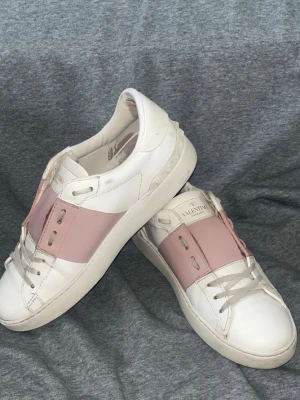 Vita sneakers med rosa detalj - Snygga vita sneakers från Valentino med bred rosa rem över mitten och coola nitar på hälen. Skorna har klassisk låg profil och vita snören. Tillverkade i läder för en lyxig känsla och stilren look.