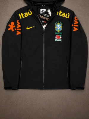 Svart Nike Brasil vindjacka - Snygg svart vindjacka från Nike med Brasilien fotbollslandslagets logga på bröstet. Jackan har huva, dragkedja framtill och färgglada tryck i gult och orange på bröst och ärmar. Tillverkad i slitstarkt material, perfekt för sportiga dagar.