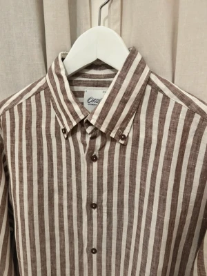 Oscar Jacobson linneskjorta - Snygg skjorta i linne med vertikala bruna och vita ränder. Klassisk krage med knappar och lång ärm. Perfekt för dig som gillar en avslappnad men stilren look. Skjortan har knäppning framtill och en rak passform. Den är i bra skick och inga defekter. Storleken är 40 vilket motsvarar ca M.