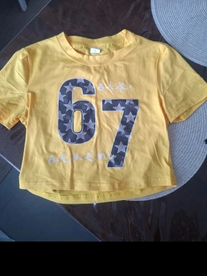 67 t-shirt med stjärnor - Gul 67  t-shirt med tryck av siffrorna 67 i grått med vita stjärnor. Texten 'six seven' står tryckt i vitt ovanför och under siffrorna. Perfekt för en avslappnad och trendig stil. Kort.