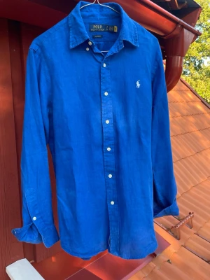 Blå linneskjorta från Polo Ralph Lauren - Säljer en klassisk blå skjorta från Polo Ralph Lauren i linne. Skjortan har lång ärm, vit broderad logga på bröstet och vita knappar framtill. Perfekt för dig som gillar en stilren och avslappnad look.