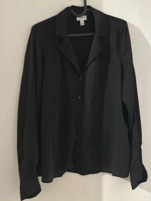 Svart långärmad skjorta i viskos - Stilren svart skjorta med lång ärm och klassisk krage. Skjortan har knappar framtill och är tillverkad i ett mjukt, lätt material som ger ett avslappnat fall. Perfekt att styla lager på lager eller bära som den är.
