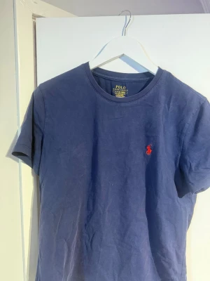 Marinblå t-shirt från Polo Ralph Lauren - Snygg marinblå t-shirt från Polo Ralph Lauren med klassisk röd logga broderad på bröstet. Modellen är slim fit och har rund halsringning samt korta ärmar. Perfekt basplagg i mjuk bomull som funkar till allt.