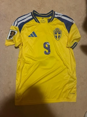 Sverige 2026 Matchtröja Herr - Alexander Isak - Säljer en officiell svensk landslagströja från Adidas med nummer 9 och namnet Isak på ryggen. Tröjan är gul med blå detaljer, har korta ärmar och FIFA VM-märken på båda ärmarna. Materialet är lätt och andas, perfekt för fotboll eller träning.