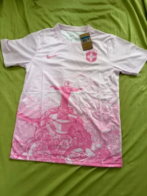Rosa Brasil fotbollströja Nike - Snygg rosa fotbollströja från Nike med Brasilien CBF-logga på bröstet och motiv av Kristusstatyn i Rio de Janeiro. Tröjan har rund hals och är tillverkad i ett lätt och ventilerande material, perfekt för träning eller match.