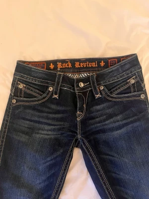 Rock Revival mörkblå jeans - Snygga mörkblå jeans från Rock Revival med kontrastsömmar och dekorativa bakfickor med broderade detaljer. Klassisk femficksmodell och låg midja. Jeansen har slitningar och tvättade effekter för en cool look. Perfekta för dig som gillar streetstyle.
