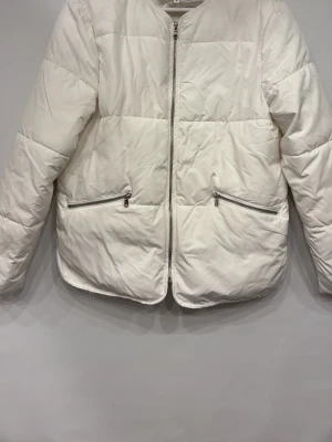Vit pufferjacka med dragkedjor - Snygg vit pufferjacka med raka linjer och quiltad design. Jackan har två framfickor med silverfärgade dragkedjor och stängs med en matchande dragkedja framtill. Perfekt för kalla vinterdagar och ger en clean, modern look.