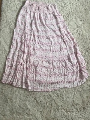 Lång blommig kjol i ljusrosa toner - Säljer en lång kjol i ljusrosa och vitt. Kjolen har smock i midjan för skön passform och är tillverkad i ett lätt, luftigt material. Jag har använt den många gånger men man ser inga tydliga tecken på användning.