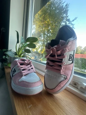 DC Shoes rosa och vita sneakers - Säljer ett par snygga DC Shoes sneakers i rosa och vitt med svarta detaljer och klassisk logga på sidan. Skorna har låga skaft, perforerad tå och rosa snören. Perfekta för dig som gillar streetwear och skate-inspirerad stil.