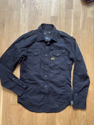 Svart Gstar skjorta med knappar - Säljer en svart långärmad skjorta med knappar framtill och två bröstfickor. Skjortan har en klassisk krage och axelklaffar för en cool touch. Passar perfekt till jeans.