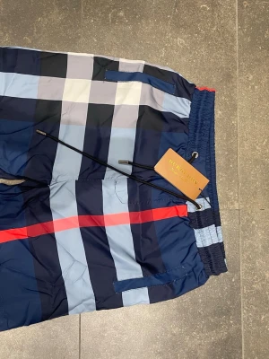 Rutiga badbyxor från Burberry - Snygga badbyxor från Burberry med klassiskt rutmönster i blått, vitt, svart och rött. De har elastisk midja med dragsko och sidofickor. Perfekta för stranden eller poolen, tillverkade i snabbtorkande material.