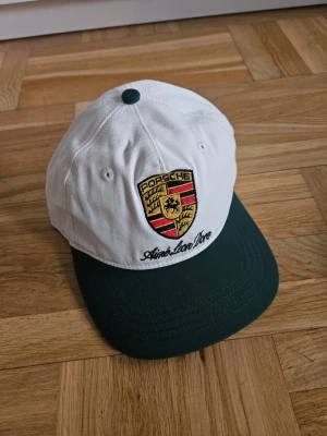 Porsche x Aime Leon Dore keps - Snygg keps från Aime Leon Dore x Porsche i vitt och mörkgrönt med klassiskt Porsche-emblem broderat framtill. Kepsen har rundad skärm och är tillverkad i bomullstyg. Perfekt accessoar för dig som gillar highend och streetwear.