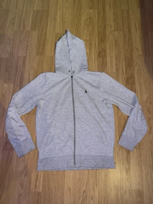 Ljusgrå zip hoodie med huva - Säljer en stilren ljusgrå Ralph Lauren hoodie med dragkedja och huva. Dragkedjan har gått sönder men går fortfarande att dra upp och ned.