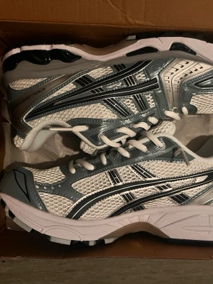 Asics gel kayano 14 fjordgrey/silver - Säljer ett par snygga Asics sneakers i vitt och grått med klassisk mesh och syntetmaterial. Skorna har tydliga Asics-loggor på sidorna och vita snören. Perfekta för dig som gillar sportig stil och vill ha bekväma skor till vardags.