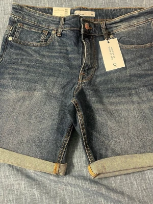 Blå jeansshorts med uppvik - Säljer ett par klassiska blå jeansshorts med uppvikta benslut och fem fickor. Shortsen har en normal passform och stängs med knapp och dragkedja. Perfekta för sommaren och lätta att matcha med olika stilar.