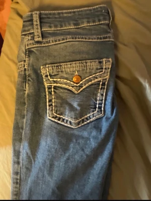 Blå jeans med kontrastsömmar - Säljer ett par blå jeans med tydliga vita kontrastsömmar och snygga detaljer på bakfickan. har ett hål på ena låret, skriv vid bild på de! 