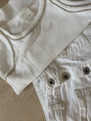 Vit ribbad croppad linnetopp och jeans kjol från ZARA - Säljer en vit ribbad linnetopp från ZARA med croppad passform. Toppen har snygga detaljer med silverfärgade sömmar längs kanterna och är ärmlös. Perfekt till sommarens outfits!