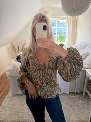 Adoore blus - Säljer en beige och brun zebramönstrad blus från Adoore med djup v-ringning och stora puffärmar. Blusen har knappar framtill och markerad midja för en snygg siluett. Kan tänka mig att byta mot en annan Adoore blus med. 