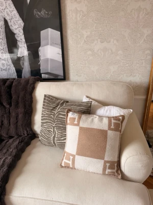 Beige och vit Hermès kudde - Säljer en lyxig kudde från Hermès i beige och vitt rutmönster med stora H-bokstäver i hörnen. Kudden har en mjuk ullkänsla och dekorativ kant. Perfekt som snygg inredningsdetalj i soffan eller sängen. lite nopprig