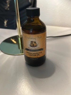 Jamaican Black Castor Oil Sunny Isle - Jamaican Black Castor Oil från Sunny Isle i en brun glasflaska med svart skruvlock. Oljan är känd för sina vårdande egenskaper för hår, ögonbryn och hud. Flaskan har en gul etikett med svart text och palmträdsmotiv.