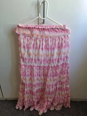 Långkjol från Gina Tricot i rosa mönster - Säljer en somrig långkjol från Gina Tricot med rosa, vita och gula toner. Kjolen har ett smockat midjeparti och volangdetaljer, samt ett färgglatt mönster som ger en bohemisk vibe. Perfekt för varma dagar och festivaler!
