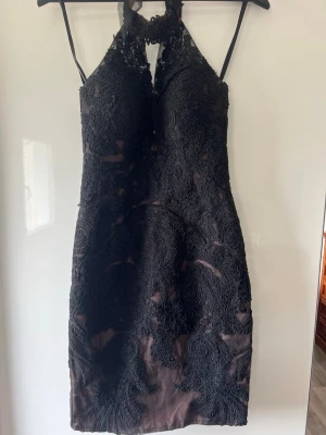 Svart spetsklänning med halterneck - Snygg svart klänning med halterneck och figurnära passform. Klänningen har ett lager av svart spets med blommönster över en beige underklänning, vilket ger en elegant kontrast. Dragkedja bak och slits nedtill. Perfekt för dig som vill sticka ut.