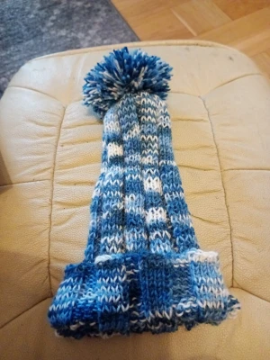 Blåvit stickad mössa med tofs one size  - Säljer en lång, stickad mössa i blått och vitt med ribbad kant och stor garnboll på toppen. Mössan har ett coolt mönster i olika blå nyanser och vitt, perfekt för kalla dagar. Passar dig som gillar en avslappnad och mysig stil. Ull,bomull polyester.tvöttråd 30 grader handtvätt 