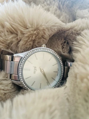 Silverfärgad klocka från Regal - Stilren klocka från Regal i silverfärg med metallarmband och en rund urtavla. Urtavlan är vit med minimalistiska markeringar och ramen är dekorerad med små stenar för extra bling. Perfekt accessoar för dig som gillar en clean look.